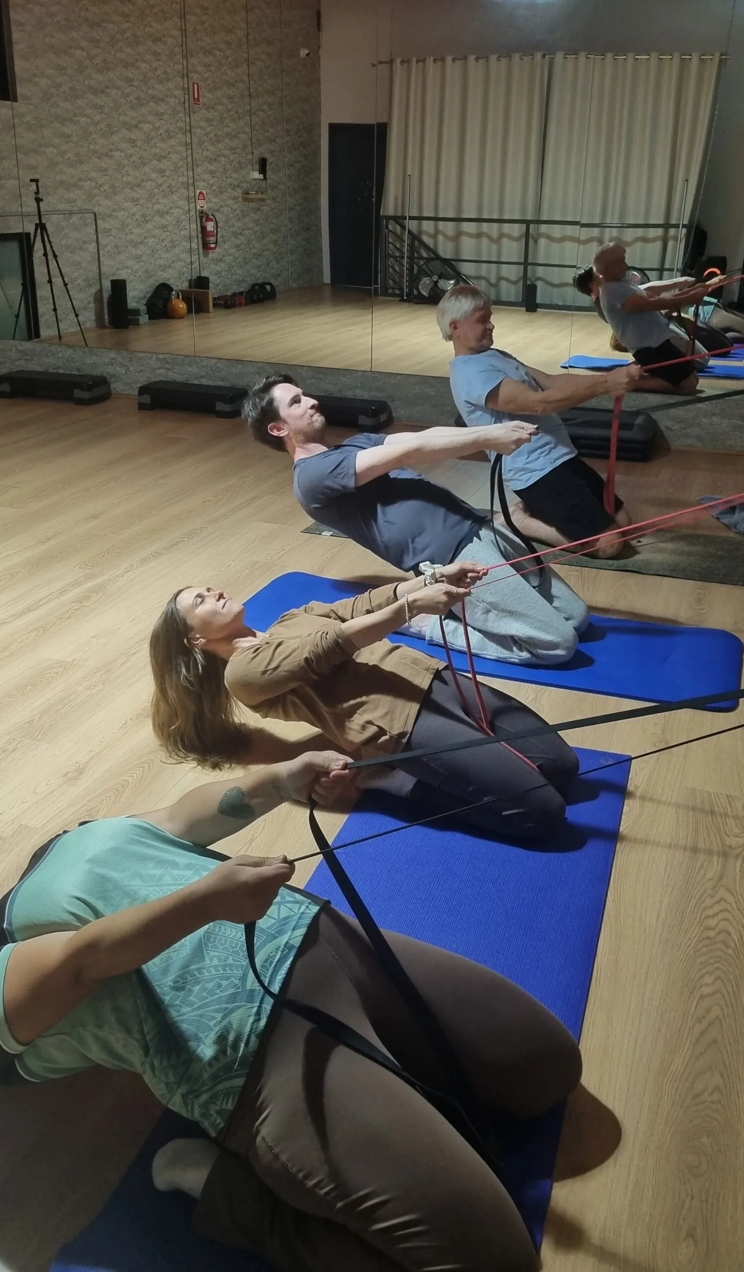 Four people kneeling on mats in a mirrored studio, leaning back and holding resistance bands overhead during a group mobility class